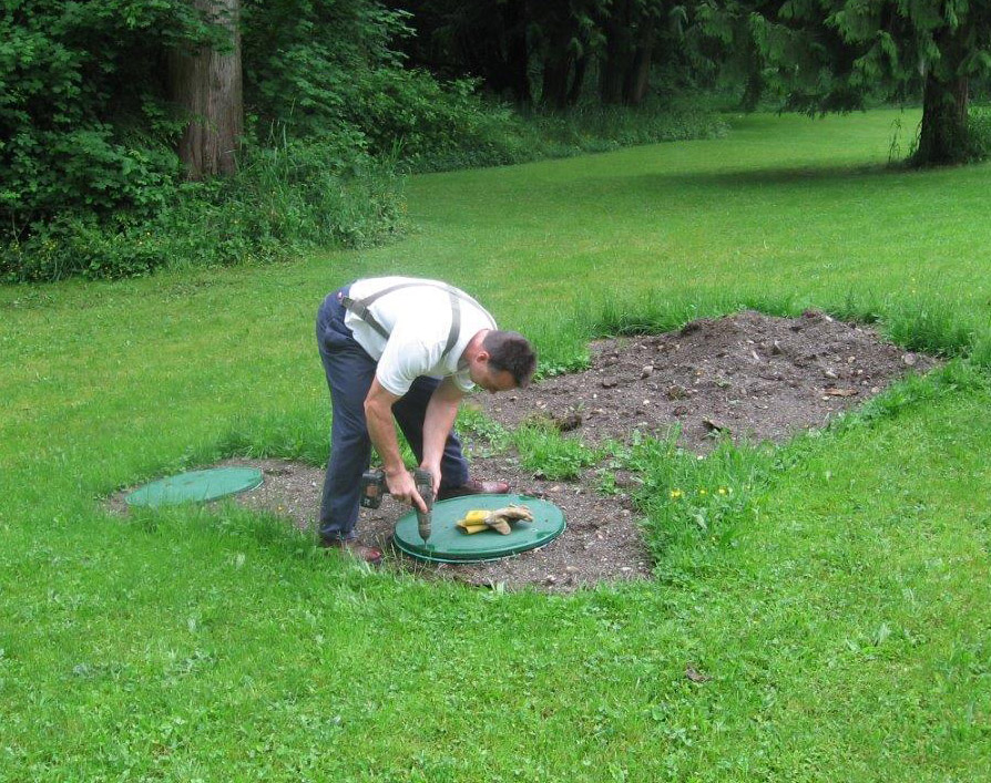 A man leans down to unscrew a green septic tank lid.