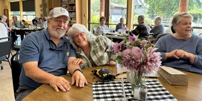 A happy couple seated at a table adorned with a flower arrangement with a friend at a senior center.