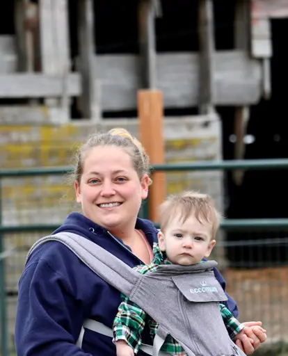 Cynthia Buchanan of Egbert Acres in Bow, WA with a young baby in a carrier on her chest and a barn in the background.